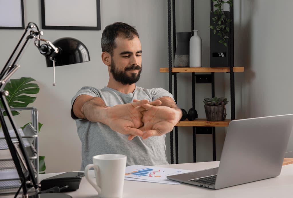 HOMBRE JOVEN EN ESCRITORIO OFICINA DISPUESTO COMIENZA A TRABAJAR SE ESTIRA LOS DEDOS DE LAS MANOS BIENESTAR TRABAJO 